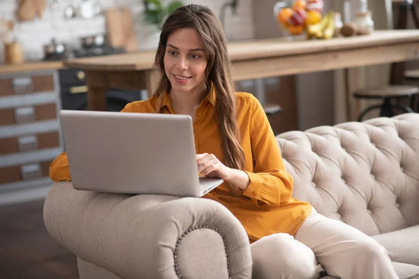 Długowłosy dziewczyna w musztardowym kolorze bluzka siedzi na kanapie z laptopem i surfing internet — Zdjęcie stockowe