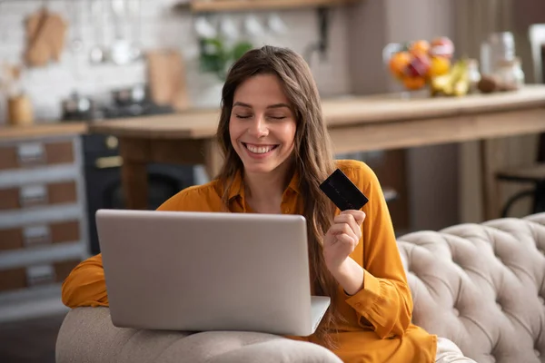 Long-haired girl in a mustard color blouse sitting on the sofa with a laptop and smiling — Stock Photo, Image
