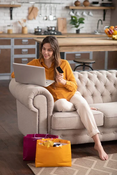 Długowłosy dziewczyna w musztardowej bluzce siedzi na kanapie z laptopem i kupuje coś online — Zdjęcie stockowe