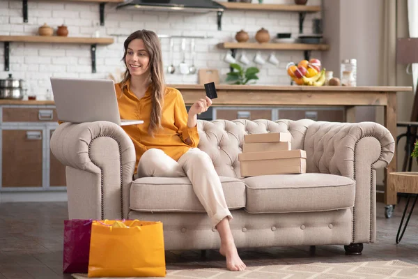 Длинноволосая девушка в горчичной блузке сидит на диване с ноутбуком — стоковое фото
