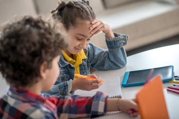 Menino e menina sentados à mesa redonda com tablets, fazendo suas atribuições, sorrindo — Fotografia de Stock