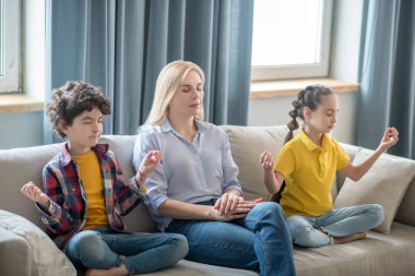 Kıvırcık çocuk, siyah saçlı kız ve sarışın kadın kanepede oturmuş meditasyon yapıyorlar.