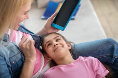 Sarışın kadın kanepede oturuyor, elinde tablet, koyu saçlı kız kucağında yatıyor, gülümsüyor.