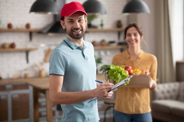 Kırmızı şapkalı genç kurye elinde alışveriş torbası ve gülümsüyor.