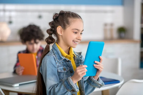 Ciemnowłosa dziewczyna trzyma laptopa, patrząc na ekran, uśmiechnięty, kręcony chłopiec siedzi przy okrągłym stole z tabletem za nią — Zdjęcie stockowe