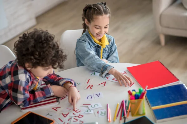 Krullenbol jongen en donkerharig meisje zitten aan ronde tafel, spelen met alfabet letters, glimlachen — Stockfoto