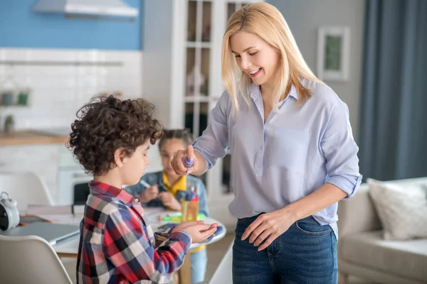 Blond vrouwtje sproeien antiseptisch op krullende jongen handen, donkerharig meisje zitten aan ronde tafel achter hen, schrijven — Stockfoto