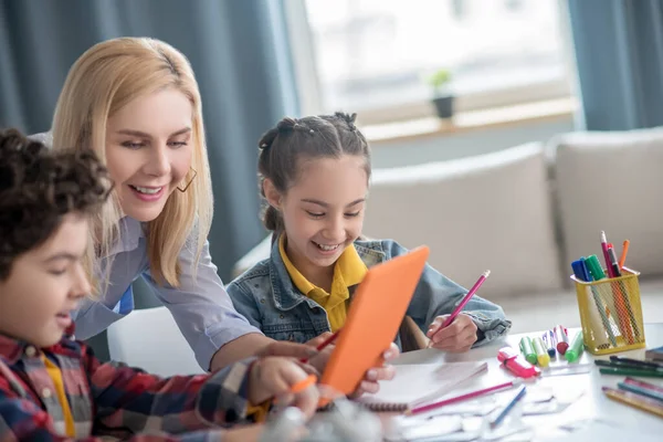 Ragazzo riccio e ragazza dai capelli scuri seduti a tavola, donna bionda che si piega tra loro, mostrando qualcosa sul tablet — Foto Stock