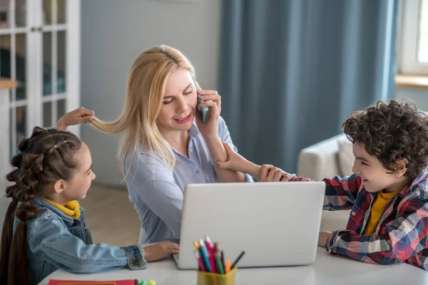 Blonde female talking on mobile and working on laptop, distracted by curly boy and dark-haired girl — Stock Photo, Image