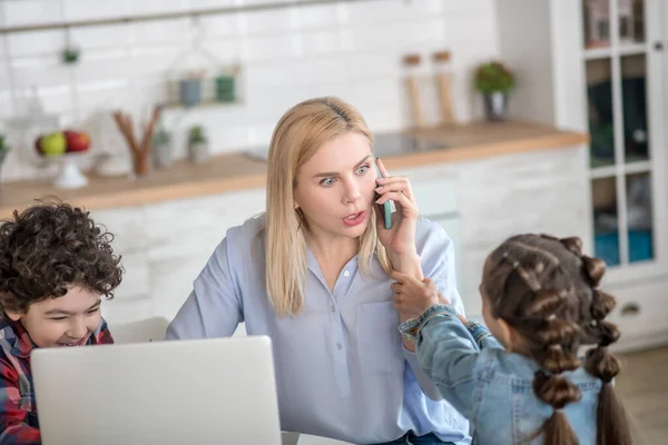 노트북으로 일하고, 옆에 앉아 있는 곱슬곱슬 한 남자 아이와 핸드폰으로 이야기하는 금발의 여성 — 스톡 사진