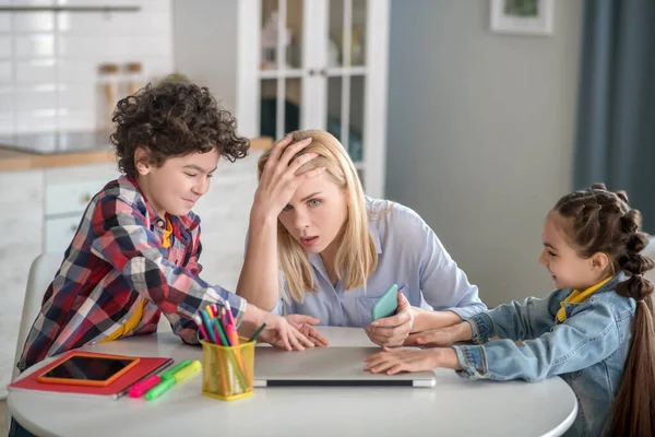 Tired blonde female sitting at table, holding her head, curly boy and dark-haired girl fighting over laptop — Stock Photo, Image