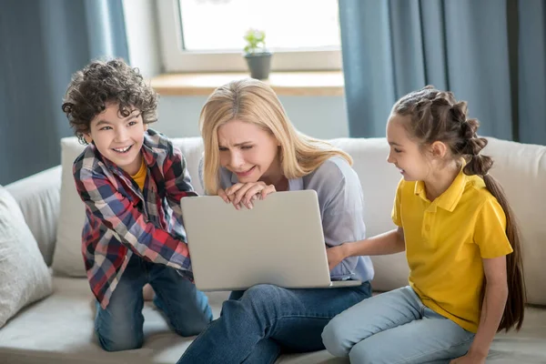 Gestresste blonde Frau sitzt auf Sofa, hält Laptop auf den Knien, lockiger Junge und dunkelhaariges Mädchen kämpfen darum — Stockfoto