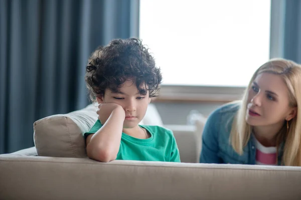 Droevige krullende jongen zit op de bank, blonde vrouw praat met hem, probeert hem op te vrolijken — Stockfoto