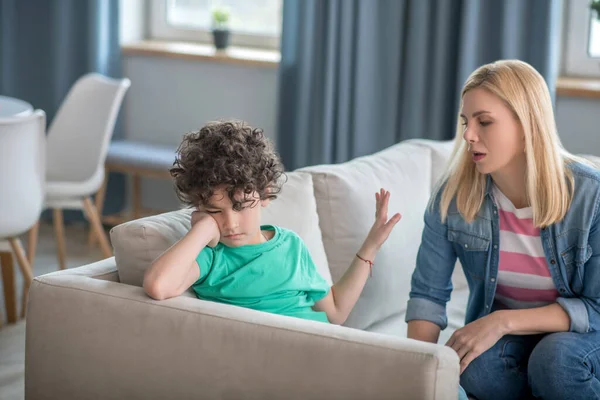 Droevige krullende jongen zit op de bank, blonde vrouw praat met hem, probeert hem op te vrolijken — Stockfoto