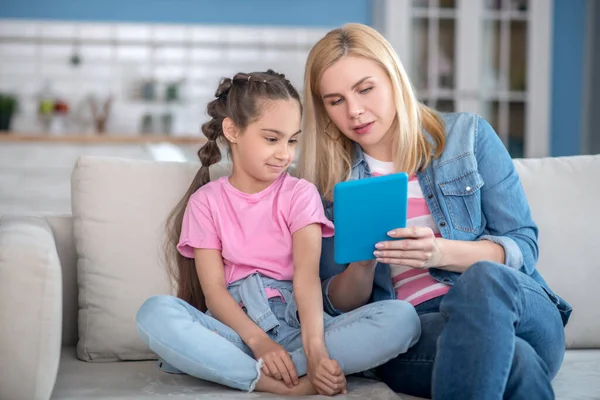 ブロンド女性と黒髪の女の子座っていますソファ,何かを見ますタブレット — ストック写真