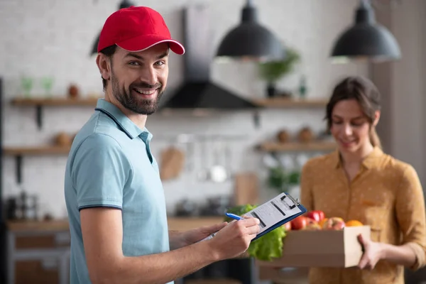 Joven cortesano en un sombrero rojo sosteniendo comestibles y sonriendo amablemente — Foto de Stock