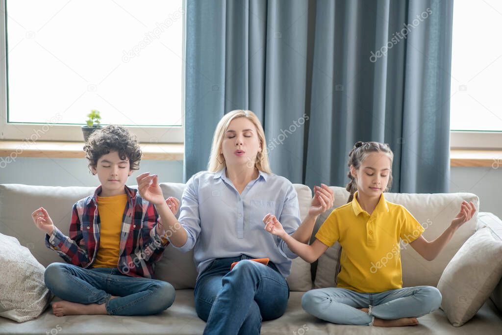 Curly boy, dark-haired girl and blonde female sitting on sofa, meditating