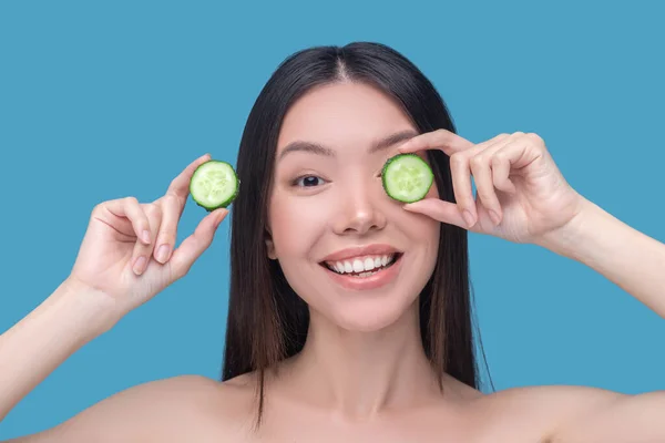 Smiling young dark-haired woman holding slices of cucumber — Stock Photo, Image
