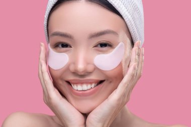 Beyaz başörtüsü takmış gülümseyen Asyalı kız gözlerinin altına yamalar yapıştırıyor.