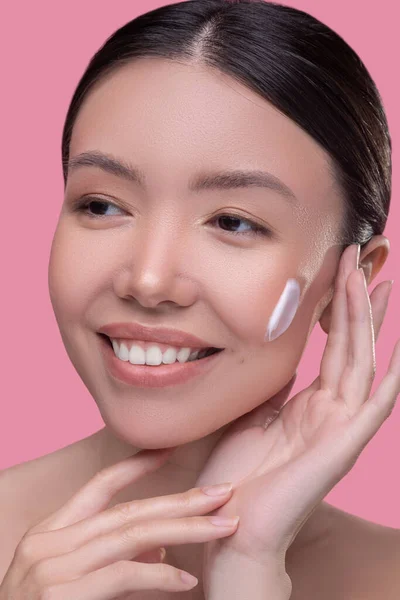 Досить азіатська дівчина наносить крем на обличчя і виглядає задоволеною — стокове фото