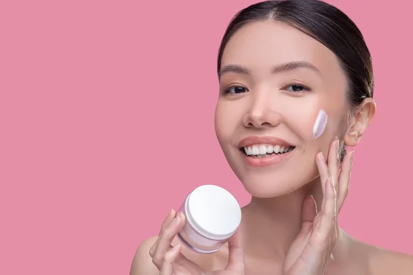 Bella ragazza asiatica applicando crema sul suo viso e sorridente — Foto Stock