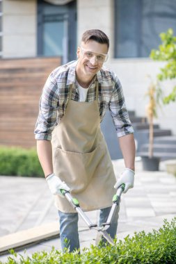 Koruyucu gözlüklü koyu saçlı erkek çalıları buduyor, gülümsüyor.