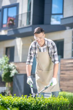 Koruyucu gözlüklü koyu saçlı erkek bahçede çalıları buduyor.