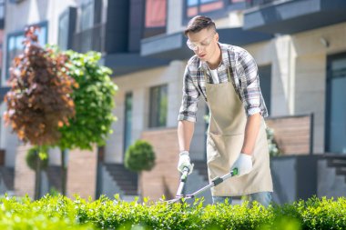 Koruyucu gözlüklü koyu saçlı erkek bahçede çalıları buduyor, gülümsüyor.