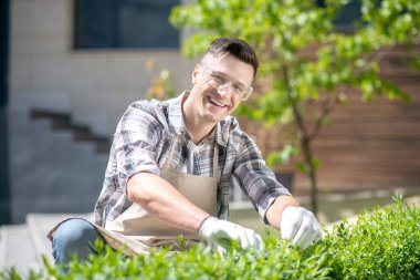 Koruyucu gözlüklü ve eldivenli neşeli siyah saçlı erkek bahçede çalıları buduyor, gülümsüyor, çömeliyor.