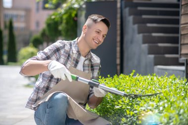 Koruyucu eldivenli koyu renk saçlı erkek dışarıda çalıları buduyor, çömelmiş.