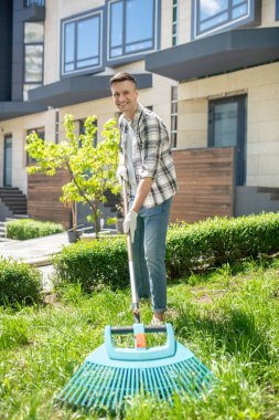 Koruyucu eldivenlerle gülümseyen siyah saçlı erkek bahçede otları tırmıklıyor.