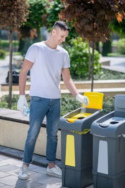 Koruyucu eldivenli siyah saçlı erkek plastik şişeleri çöp konteynırına atıyor.