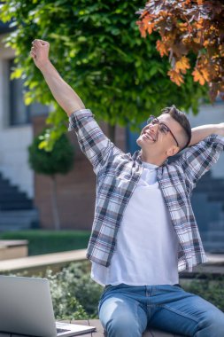 Koyu renk saçlı erkek, gözlüklü, dışarıda bankta dizüstü bilgisayarıyla oturuyor, sırtını uzatıyor.