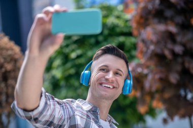 Neşeli siyah saçlı erkek kulaklık takıyor, selfie çekiyor, gülümsüyor.