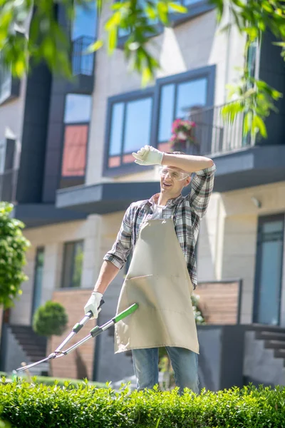 Siyah saçlı, koruyucu gözlüklü bir erkek bahçede çalıları buduyor, kaşlarını paspaslıyor.