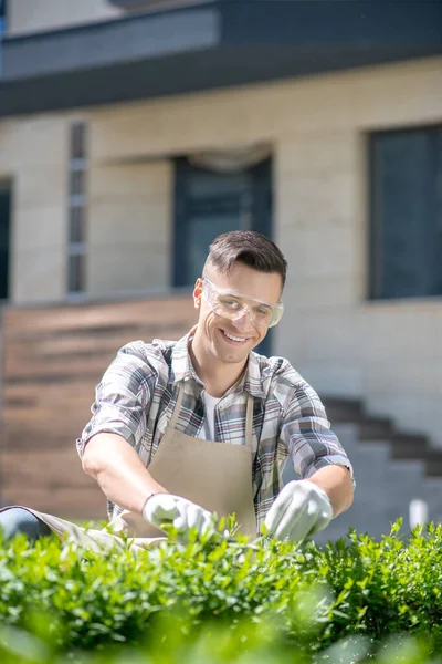 Koruyucu gözlüklü ve eldivenli koyu renk saçlı erkek bahçede çalıları buduyor, gülümsüyor.