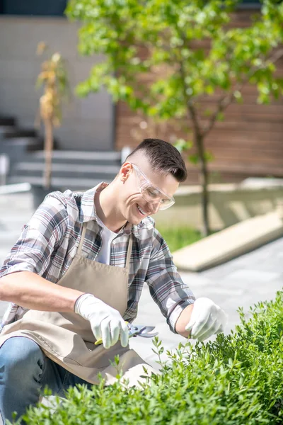 Koruyucu gözlüklü ve eldivenli neşeli siyah saçlı erkek dışarıda çalıları buduyor, gülümsüyor, çömeliyor.