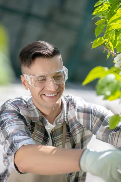 Koruyucu gözlüklü koyu renk saçlı bir erkek ve bahçede ağacı buduyor, gülümsüyor.