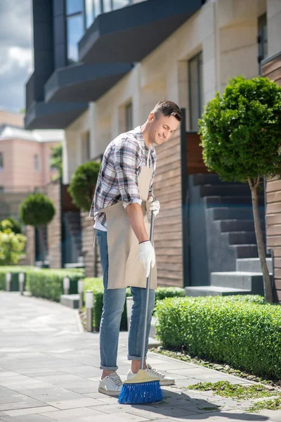 Dark-haired male in protective gloves and apron sweeping leaves in the yard — Stock Photo, Image