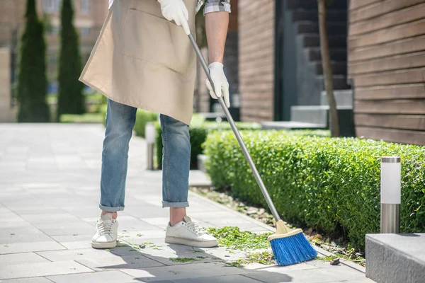 Close-up of male in protective gloves and apron sweeping leaves with broom outside — Stock Photo, Image