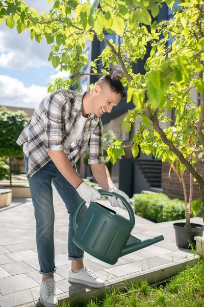 Smiling dark-haired male in protective gloves watering tree with green watering can in the yard — Stock Photo, Image