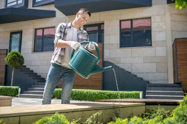 Siyah saçlı, koruyucu eldivenli, gülümseyen bir erkek bahçedeki çalıları sulayan bir teneke kutu.