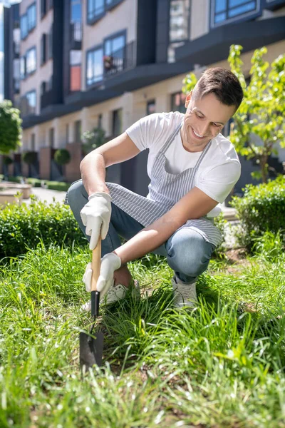 Glimlachende donkerharige man in beschermende handschoenen en schort hurkend, harkend gras in de tuin — Stockfoto