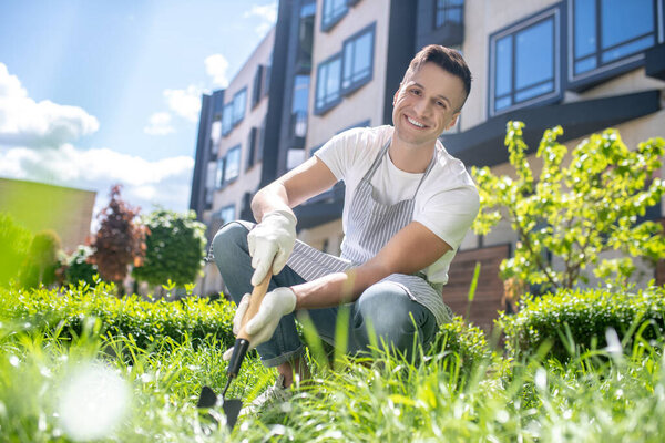 Cheerful dark-haired male in protective gloves and apron crouching, raking grass in the yard