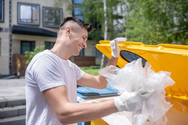 Koruyucu eldivenlerle sırıtan siyah saçlı bir erkek çöp kutusuna çöp atıyor.
