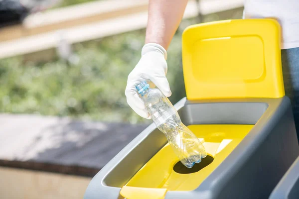 Primer plano de las manos masculinas en guantes protectores lanzando una botella de plástico abierta en un contenedor de residuos — Foto de Stock