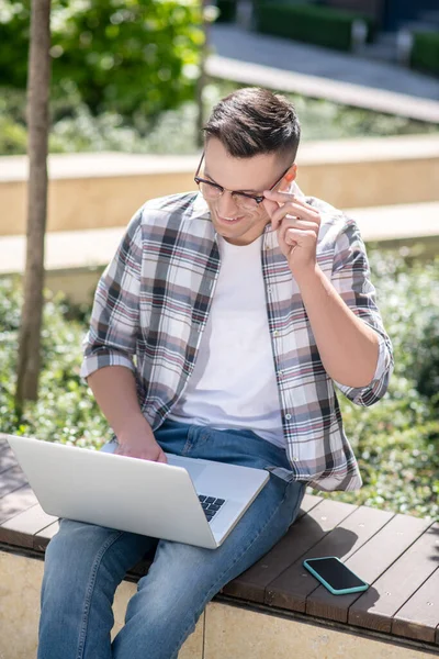 Koyu renk saçlı erkek bahçede oturuyor ve dizüstü bilgisayarıyla çalışıyor. Tek eliyle gözlüklerine dokunuyor.