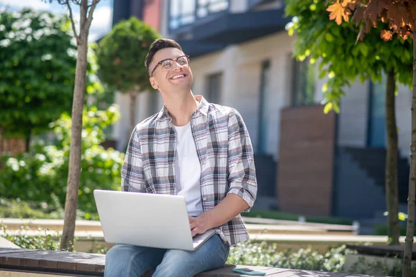 Koyu renk saçlı, gözlüklü bir erkek bahçede dizüstü bilgisayarıyla oturuyor ve gökyüzüne bakıyor.
