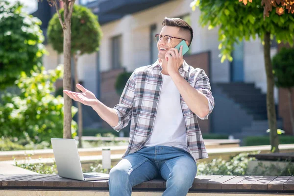 Hombre moreno con gafas, sentado en el patio con su portátil, hablando en su móvil —  Fotos de Stock