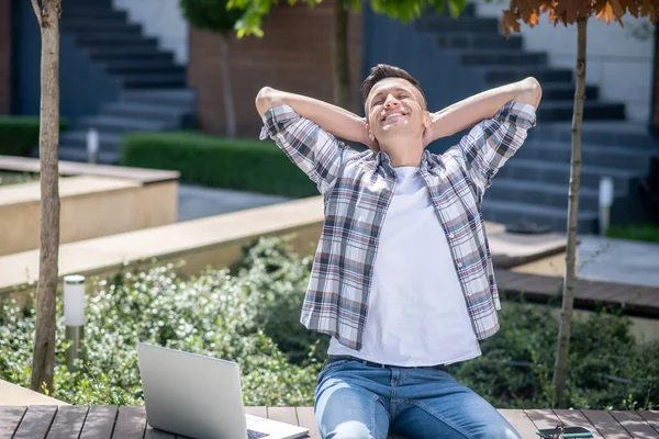 Koyu renk saçlı erkek dışarıda bankta dizüstü bilgisayarıyla oturuyor, sırtını esnetiyor.